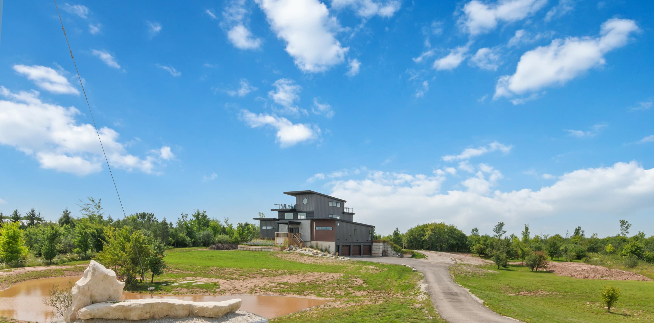 A wide view across the pond and the entrance drive to the house standing on a rise under a broad sky