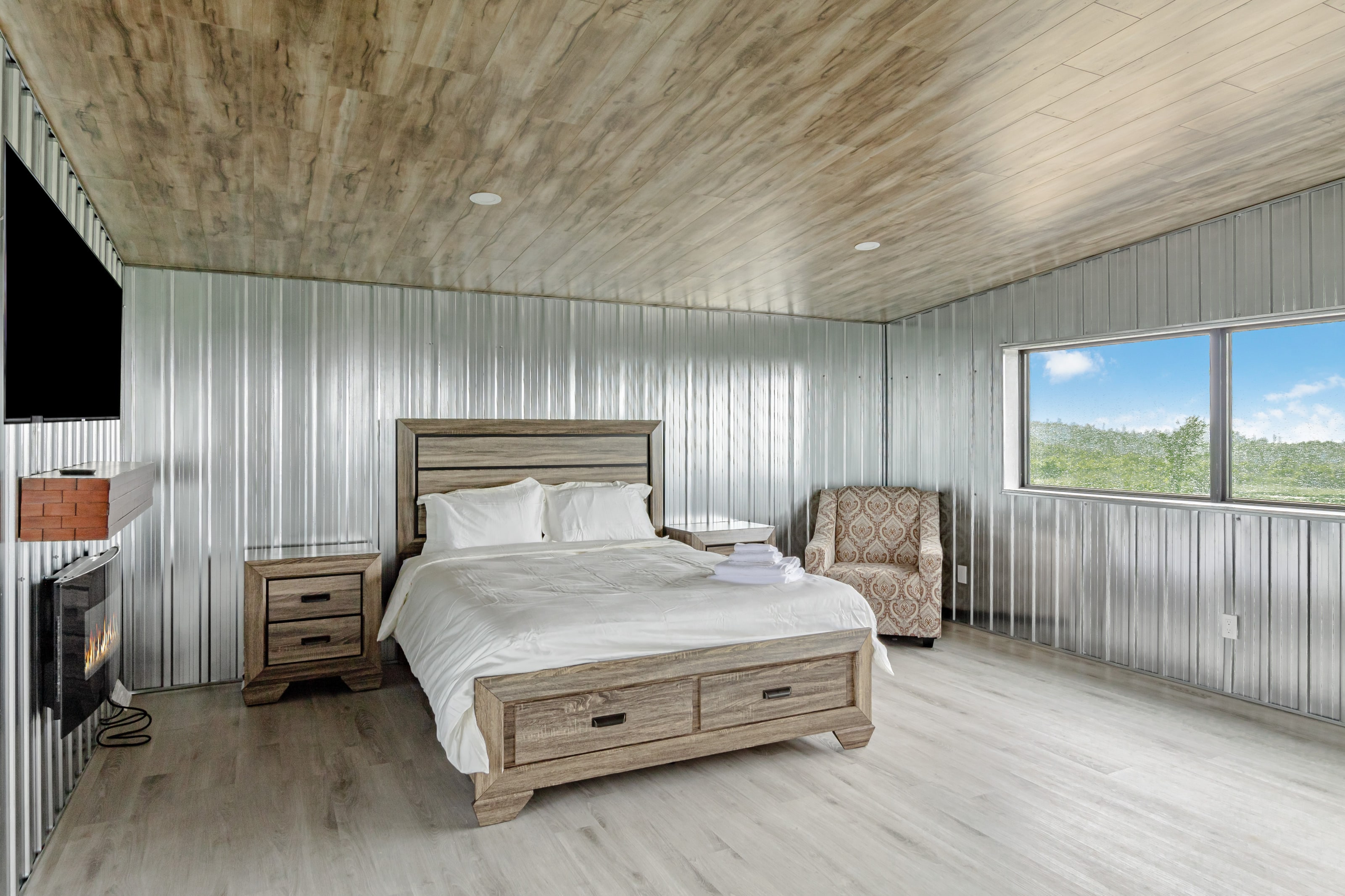 A steel-lined bedroom with a lit fireplace beside the bed and a window onto the fields