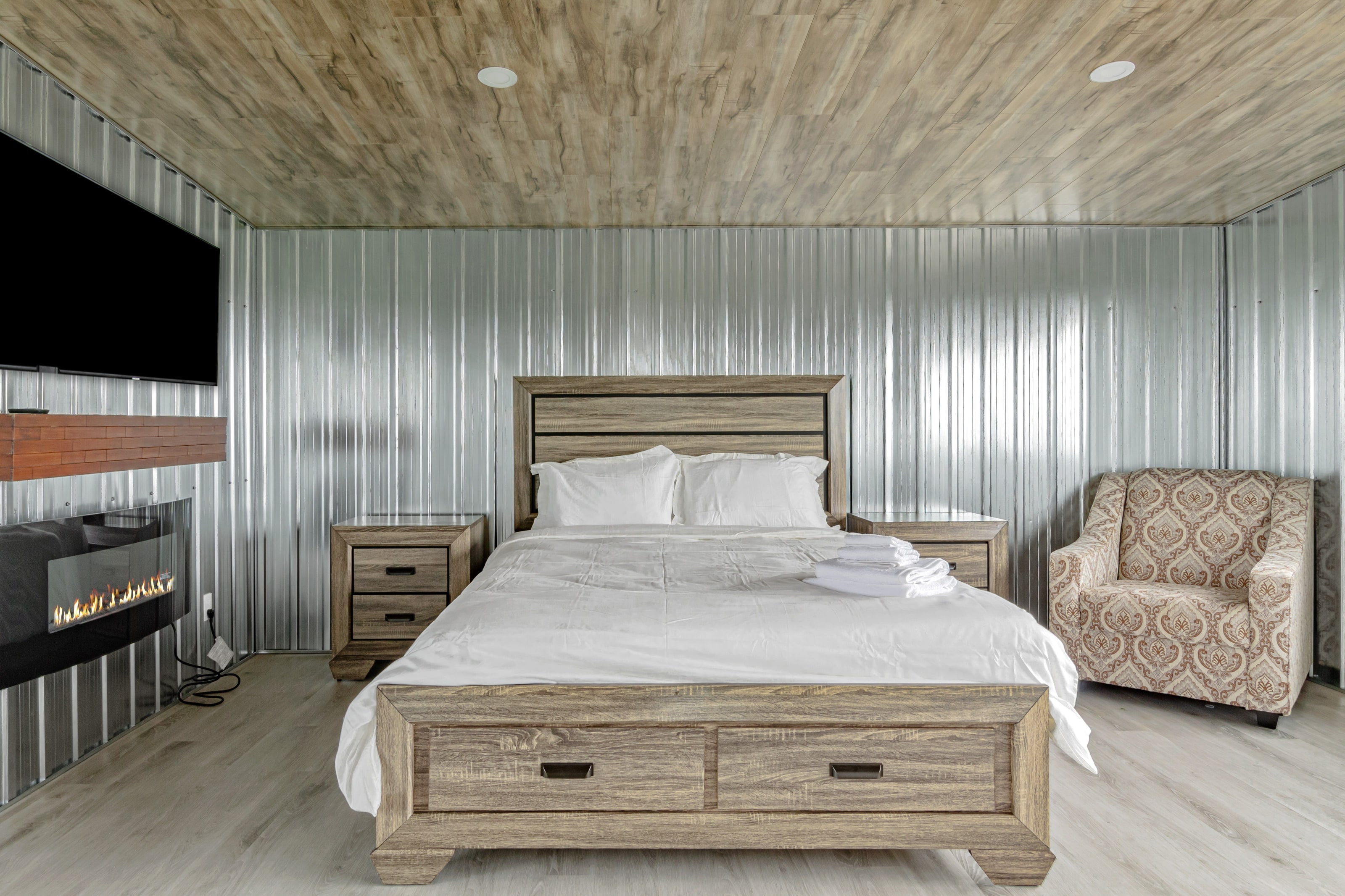 A bedroom lined in corrugated galvanised steel with a fireplace, television and timber mantel opposite the bed