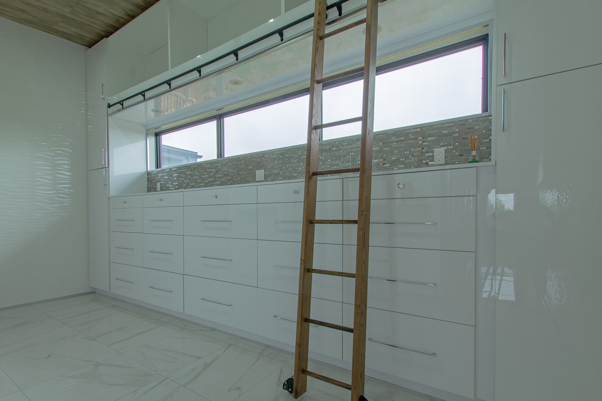 A dressing room walled in white cabinetry with a rolling timber library ladder and a ribbon window above