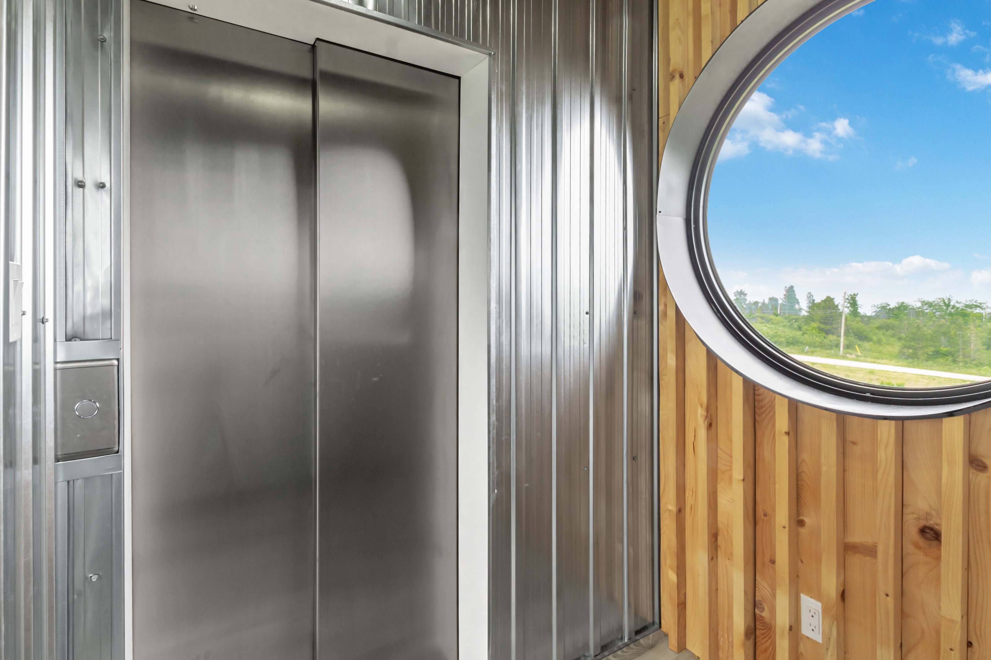 Stainless steel elevator doors set into a corrugated galvanised steel wall, beside a round porthole window in cedar planking