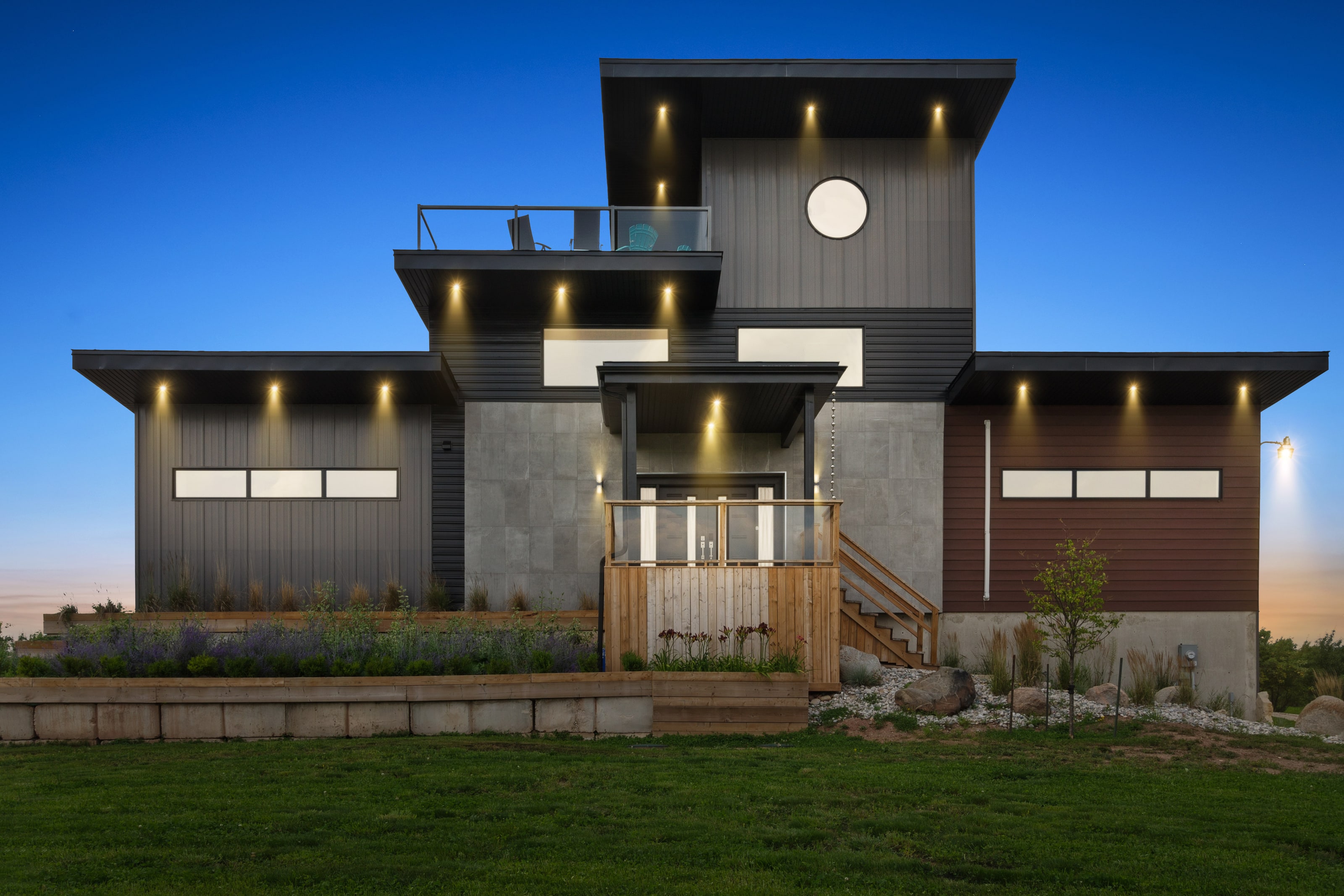 The full front elevation at dusk: stacked charcoal steel and concrete volumes lit by warm soffit lights, with a round porthole window and a rooftop terrace above