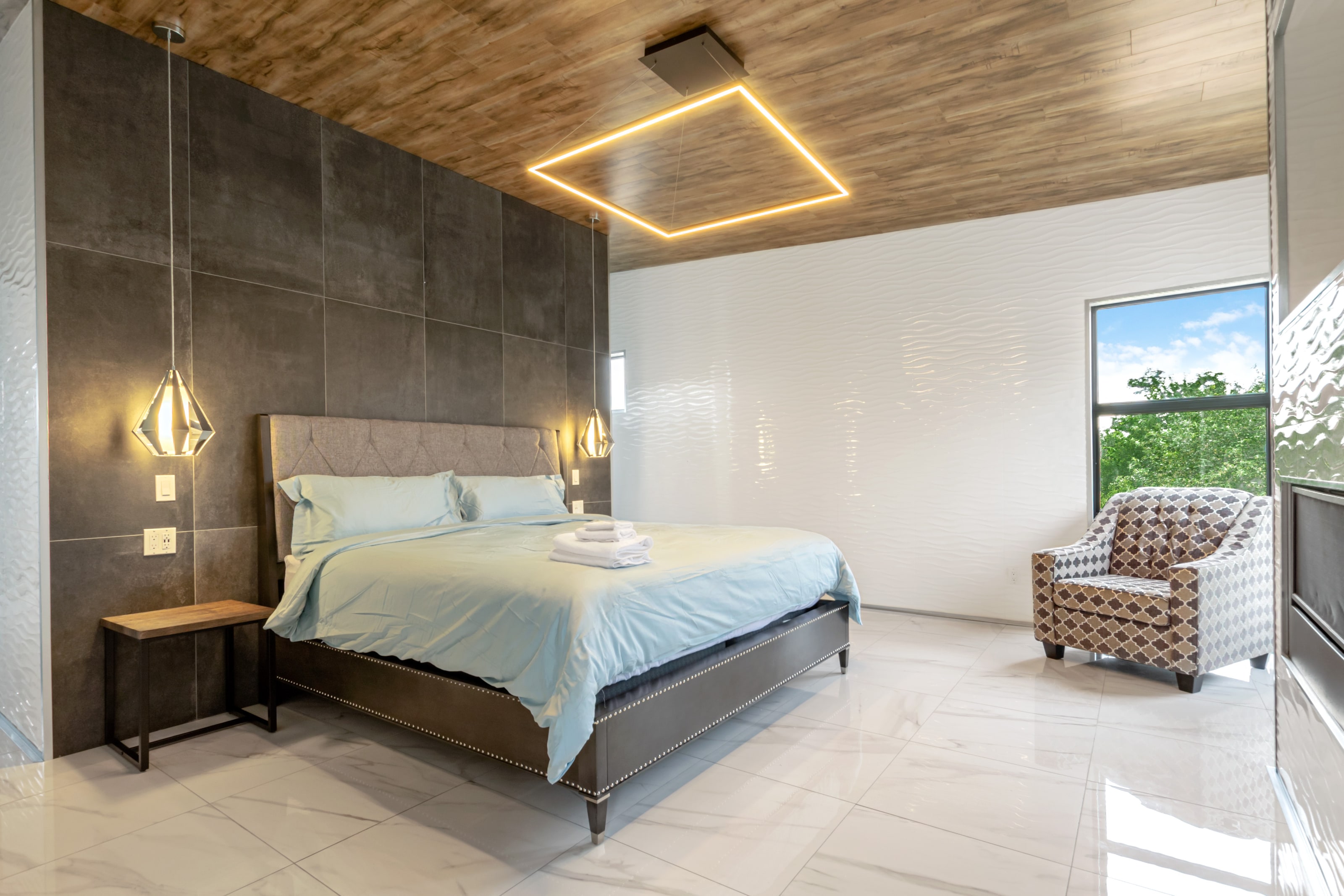 A king bed against a dark tiled feature wall, under a timber ceiling with a recessed light cove